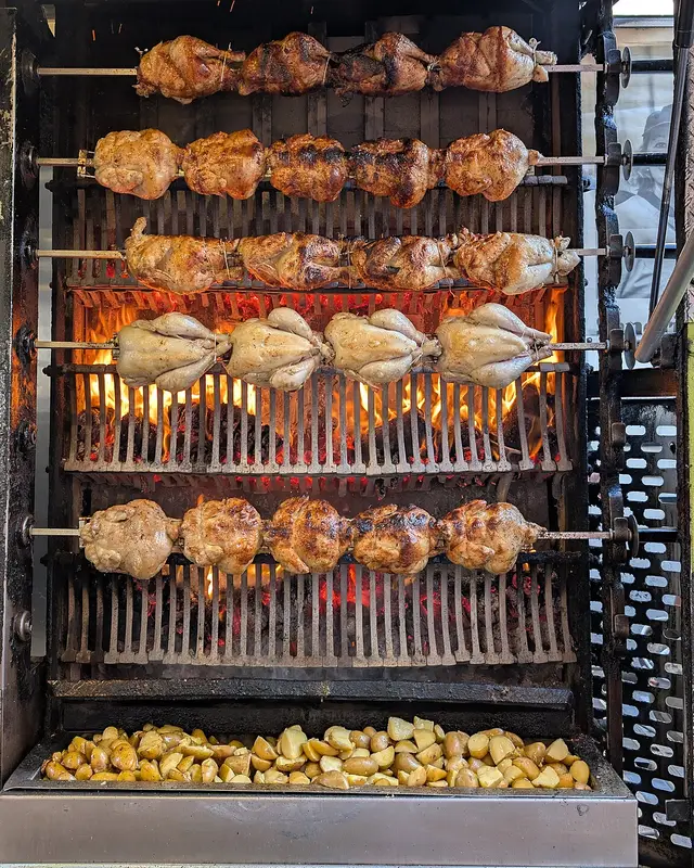 Cinco filas de pollos girando en el ast sobre las brasas, con una bandeja de patatas debajo recogiendo el jugo
